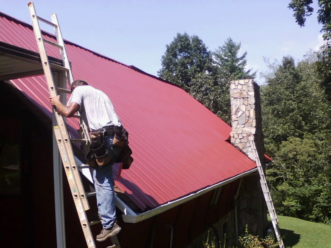 Roofer on a ladder beside a red metal roof and stone chimney — Soffit & Fascia Repair by Roofing Brothers in Teterboro