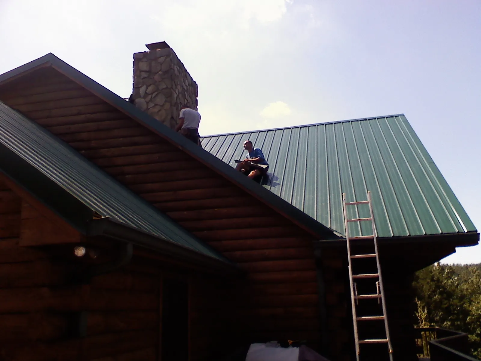 Roofers installing green metal panels beside a stone chimney on a log home in Teterboro